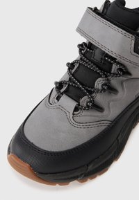 Chaussure de randonnée grise avec des accents noirs, dotée d'un collier de cheville rembourré, d'un capuchon en caoutchouc pour les orteils et d'un système de laçage flexible. Matériaux texturés utilisés.