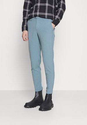 Person trägt maßgeschneiderte hellblaue Hosen, schwarze Stiefeletten mit dicker Sohle und ein schwarzes kariertes Langarmhemd, hat eine Hand in der Tasche.