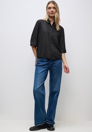 Camicia nera con maniche a tre quarti, abbinata a jeans a gamba larga blu e stivali alla caviglia neri, su uno sfondo neutro.