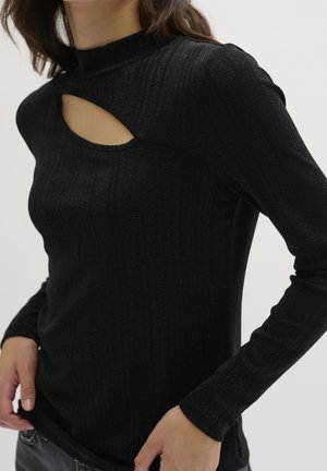 Frau trägt ein eng anliegendes, schwarz geripptes Langarmshirt mit schrägem Brustausschnitt und hohem Kragen, Hände auf den Hüften ruhend.