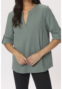 Blusa verde, con texture, scollo a V e maniche arrotolate, abbinata a pantaloni neri. Il tessuto appare leggero e dal design informale.