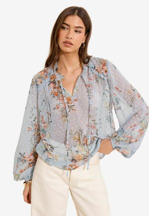 Sieviete valkā caurspīdīgu zilu blūzi ar ziedu rakstu un bīskapa piedurknēm, kopā ar augsta vidukļa krēmkrāsas biksēm un zelta lokveida auskariem.