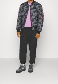 Uomo che indossa un giubbotto puffer mimetico nero e grigio, una maglietta viola, pantaloni neri Nike, calzini bianchi e sneakers grigie Nike mentre si trova su un pavimento grigio.