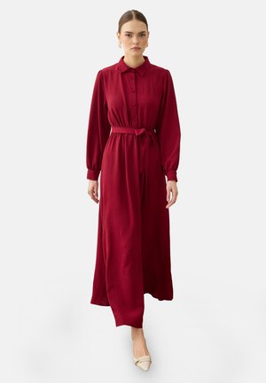 Kvinde iført en langærmet burgundy kjole med knapper, et bundet bælte i taljen og beige spidse sko på en ensfarvet baggrund.