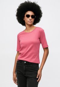 Γυναίκα με αφρικανική κοτσίδα που φορά καφέ γυαλιά ηλίου, ροζ μπλούζα κοντού μανικιού και μαύρα ψηλόμεσα τζιν μπροστά από ένα απλό φόντο.