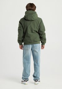 Boy standing facing away wearing an olive green hooded jacket, light blue jeans, and white Nike sneakers on a plain background.