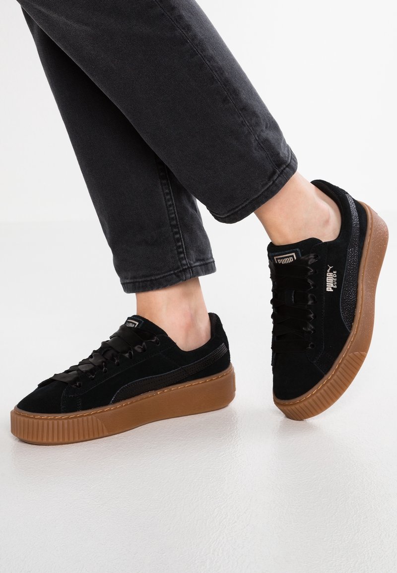 Personne portant des baskets en daim noires avec des semelles épaisses marron et des lacets noirs, associées à un pantalon gris foncé retroussé, debout sur un sol blanc.