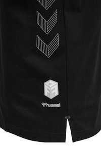 Camiseta negra de algodón con patrones gráficos blancos, que incluyen chevrones y logotipos, con una pequeña abertura lateral en el dobladillo.