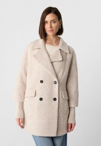 Beige Doppelreiher-Mantel aus strukturiertem Stoff. Verfügt über große Revers, zwei frontale Taschen und schwarze Knöpfe; mit gerippten Bündchen abgerundet.