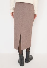 Personne portant une jupe midi en tricot marron avec une fente arrière, des collants noirs, des bottines noires et un pull crème, vue de derrière.