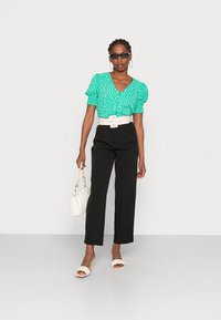 Blusa verde com bolinhas, com mangas curtas e franzidas, cinto largo branco, calças pretas de cintura alta e sandálias de salto claras.