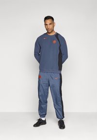 Tuta sportiva blu navy con logo Nike arancione, composto da una felpa con collo a giro e pantaloni affusolati con polsini alla caviglia e dettagli laterali.