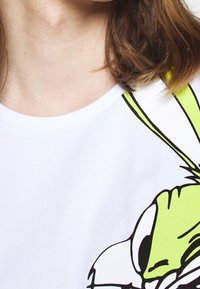 Detailní záběr na osobu nosící bílý tričko s velkým zeleným kresleným obličejem králíka na pravé straně.