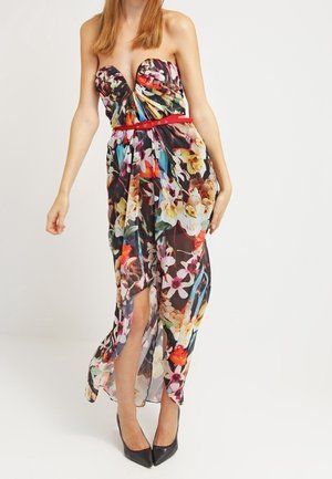 Femme portant une robe maxi sans bretelles à fleurs colorées avec une ceinture rouge et des talons hauts noirs, debout devant un fond blanc.