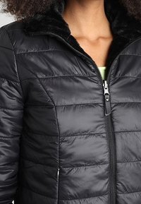 Svart puffjacka med horisontell quiltning och en mjuk, svart syntetpälskrage. Har en framdragkedja och sidofickor.