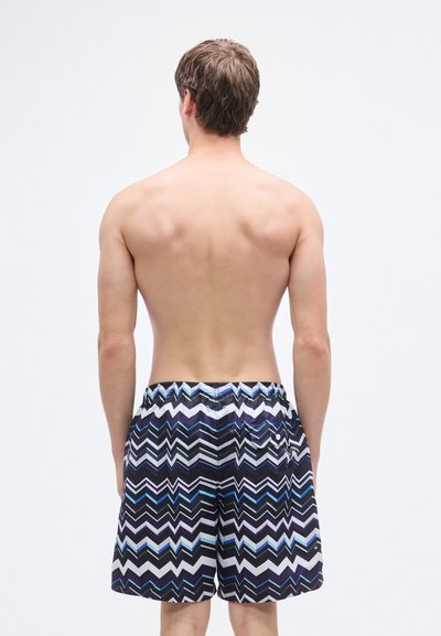 Μαγιό βερμούδα με ζιγκ-ζαγκ σχέδιο σε μαύρο, λευκό και μπλε χρώμα, με ελαστική μέση και πλαϊνές τσέπες, κατασκευασμένες από ελαφρύ ύφασμα.