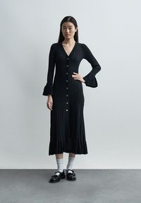 Μια γυναίκα στέκεται μπροστά σε μονόχρωμο τοίχο, φορώντας ένα μαύρο φόρεμα με κουμπιά και φουσκωτά μανίκια, λευκές κάλτσες και μαύρα μοκασίνια, με το χέρι στη μέση.