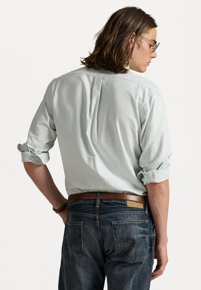 Chemise à boutons rayée vert clair avec des manches roulées, portée avec un jean bleu foncé et une ceinture marron. Texture du tissu visible et détails de couture.