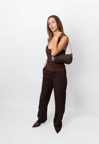 Rjava majica brez rokavov z nabranim materialom, temne hlače in čevlji z ostrimi konicami. Model drži majhno rjavo teksturirano torbico s paščkom za ramo.