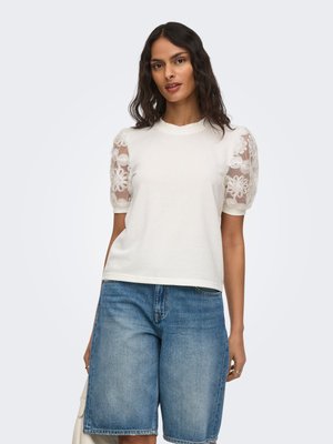 Kvinde iført hvid top med gennemsigtige blomstrede pufærmer og blå denimshorts i mellemlang længde, stående foran en ensfarvet lyse baggrund.