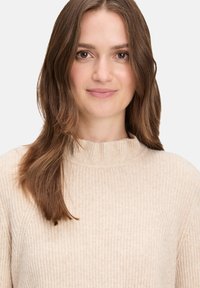 Beżowy sweter z prążkowanej tkaniny i wysokim kołnierzem. Miękka, strukturalna tkanina i dopasowany krój. Modelka ma średniej długości brązowe włosy, na jasnym tle.