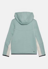 Kapucňová bunda v svetlo modro-zelenom materiáli, s krémovými bočnými panelmi a tmavým lemovaním. Hladká textúra s priliehavým strihom a detailom loga Nike.