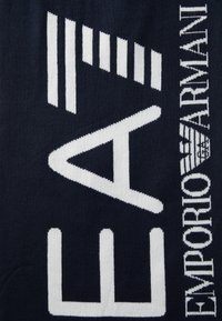 Textil de culoare bleu marin cu litere albe proeminente, inscripționat cu "EA" și "Emporio Armani." Materialul pare moale, cu o textură țesută.