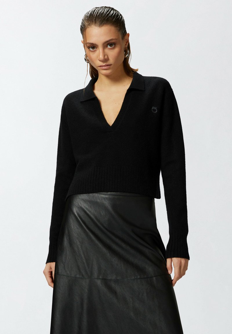 Crni skraćeni pulover s V-izrezom i rebrastim rubom, u kombinaciji s crnom kožnom suknjom koja ima glatku teksturu i detalje šavova.