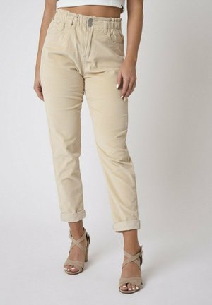 Pantalons en velours côtelé beige avec une taille élastique, bouton avant, deux poches et ourlets roulés, associés à des talons hauts beiges à lanières.
