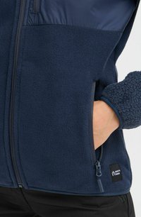 Veste en polaire bleu marine avec une partie supérieure lisse, une partie inférieure texturée, une poche zippée et une étiquette de marque à la taille.