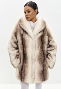 Cappotto in pelliccia sintetica di colore marrone chiaro e crema con una texture morbida, colletto oversize e tasche anteriori. Il modello indossa una camicia bianca sotto.