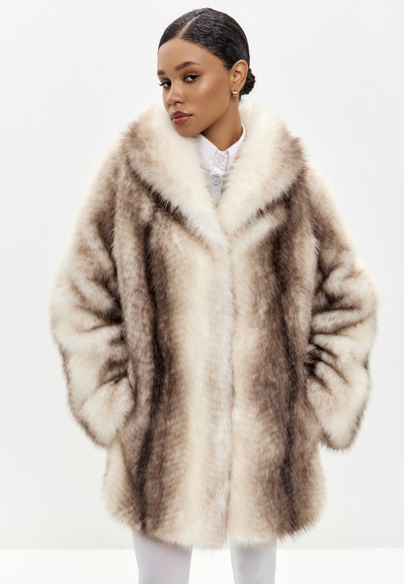 Cappotto in pelliccia sintetica di colore marrone chiaro e crema con una texture morbida, colletto oversize e tasche anteriori. Il modello indossa una camicia bianca sotto.