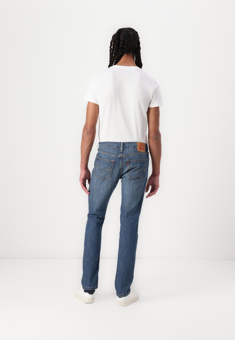 T-shirt blanca ajustada combinada con vaqueros de mezclilla azules, que presentan un corte clásico, diseño de cinco bolsillos y parche de cuero con marca en la parte trasera.