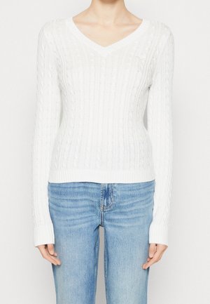 Woman wearing a white V-neck cable knit sweater and light blue denim jeans, shown from neck to hips on a plain background.