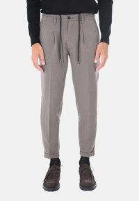 Pantaloni grigi su misura con coulisse e chiusura a bottone, dotati di pieghe frontali e orli risvoltati. Indossati con scarpe scure e calzini.