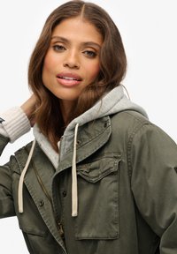 Parka verde oliva con una sudadera gris con capucha debajo. Presenta un cierre de cremallera, bolsillos laterales y un detalle de puños acanalados.