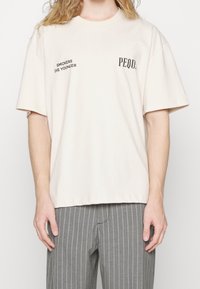 Beżowa bawełniana koszulka z krótkim rękawem, okrągłym dekoltem, z czarnym napisem: "PALACZE UMIERAJĄ MŁODZIE" oraz "PEQUUS" z przodu. Luźny fason.