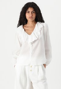 Blusa bianca con colletto arricciato, apertura frontale con bottoni e maniche lunghe. Realizzata in tessuto leggero con una texture leggermente trasparente.