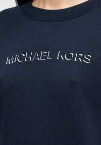 Μπλε ναυτικό φούτερ με στρογγυλή λαιμόκοψη· διαθέτει ανάγλυφο λογότυπο "MICHAEL KORS" σε λευκό χρώμα στο στήθος· κατασκευασμένο από μαλακό, υφασμάτινο ύφασμα με υφή.