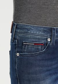 Los vaqueros de mezclilla azul oscuro presentan un diseño clásico de cinco bolsillos, con un pequeño bolsillo acentuado por rayas rojas y blancas. Se pueden ver detalles de metal plateado.