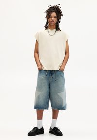 Beige mouwloze shirt, oversized pasvorm; lichtblauwe denimshorts met sterpatronen; witte sokken; zwarte schoenen; casual stijl.