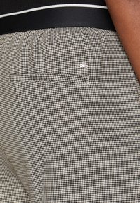 Musta-valkoiset ruudulliset housut, joissa on teksturoitu kangas, takatasku ja pieni brändilabel sivussa.