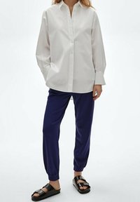 Chemise blanche à manches longues avec boutons, pantalon bleu marine avec poignets élastiques, et sandales noires à glissière avec deux larges brides.