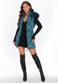 Turkusowa kamizelka ocieplana z teksturowanym, pikowanym wzorem oraz kapturem z sztucznego futra. Posiada kieszenie na zamek błyskawiczny oraz pełny zamek z przodu. W zestawie z czarną sukienką i kozakami za kolano.