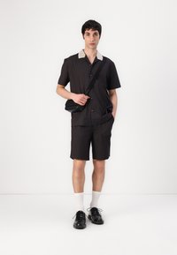 Chemise à manches courtes à rayures noires avec un col beige, associée à un short noir jusqu'aux genoux. Portée avec des chaussures noires et des chaussettes blanches, portant un sac noir.