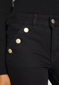 Jean noir avec un design ajusté, comportant quatre boutons dorés et une texture lisse, rehaussée de fines coutures.