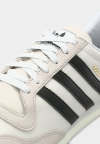 Hvit sneakers med overdel i lerret, grå semskede detaljer, tre svarte striper og en logo på tungen, mot en enkel bakgrunn.