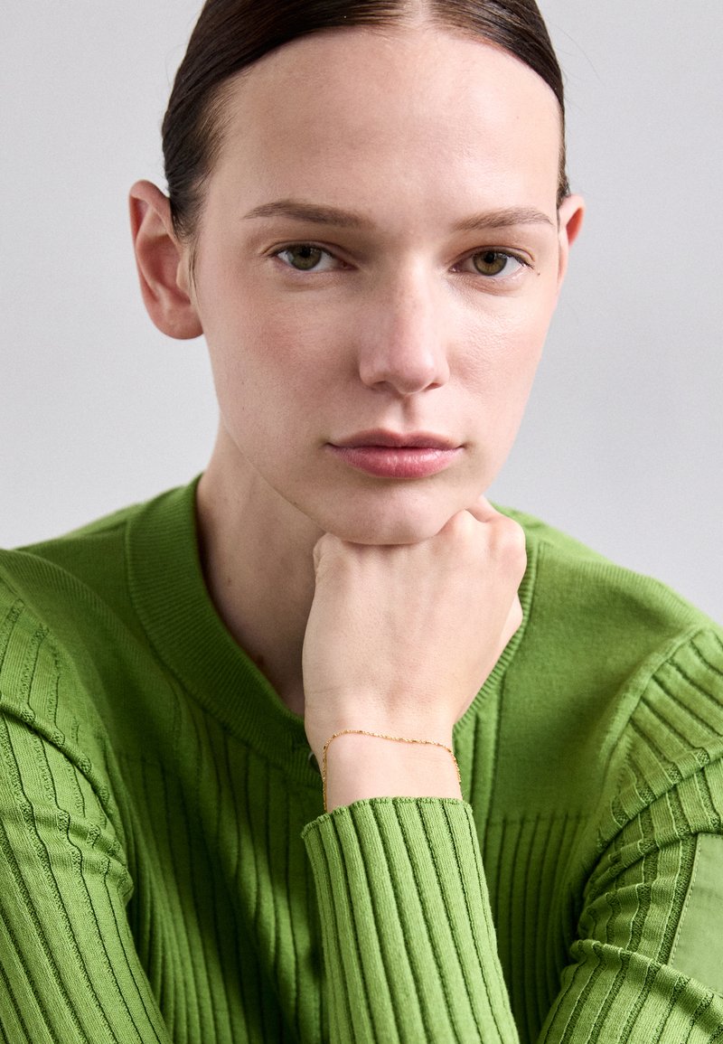 Zeleno rebrasto pleteno pulover z visokim ovratnikom, ki prikazuje teksturirane vzorce; zlat zapestnica na zapestju dodaja rahlo noto.