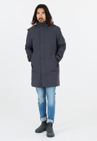 Donkergrijze parka met hoge kraag, zijzakken, lange mouwen en een rechte snit, gecombineerd met lichtblauwe jeans en zwarte enkellaarzen.