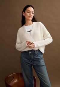  ivoorkleurige geribbelde gebreide cardigan met een sjawl-kraag, gecombineerd met lichtblauwe recht-pas jeans. Bruine stoel op de voorgrond. Neutrale achtergrond.